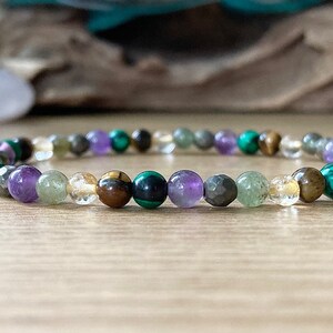 Pu&ograve; includere: Un braccialetto di perline con una variet&agrave; di pietre preziose colorate. Il braccialetto include perline verdi, viola, gialle e marroni. Le perline sono infilate su un cordino elastico trasparente. Il braccialetto &egrave; esposto su una superficie di legno.
