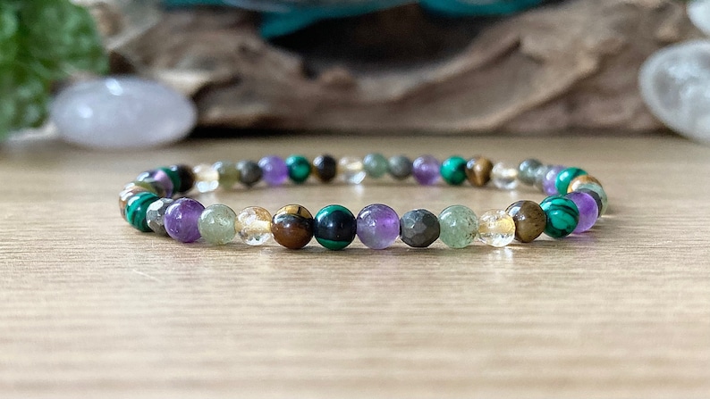 Pu&ograve; includere: Un braccialetto di perline con una variet&agrave; di pietre preziose colorate. Il braccialetto include perline verdi, viola, gialle e marroni. Le perline sono infilate su un cordino elastico trasparente. Il braccialetto &egrave; esposto su una superficie di legno.