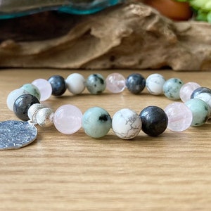 Puede incluir: Un brazalete hecho con cuentas verdes, blancas y rosas con un dije de plata. El brazalete est&aacute; sobre una superficie de madera.