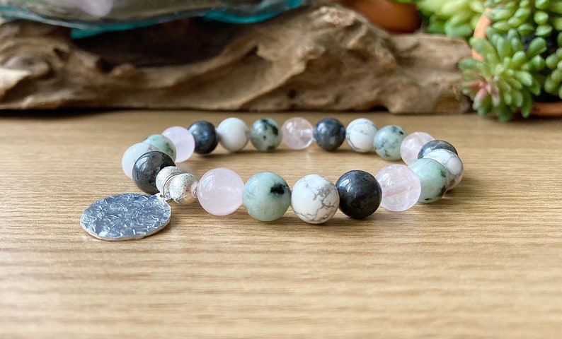 Puede incluir: Un brazalete hecho con cuentas verdes, blancas y rosas con un dije de plata. El brazalete est&aacute; sobre una superficie de madera.