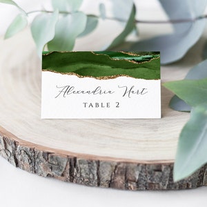 May include: A white place card with a watercolor design of dark green and gold glitter accents. The card features the name "Alexandria Hart" and "TABLE 2" in elegant script. The card is displayed on a wooden slice with greenery.