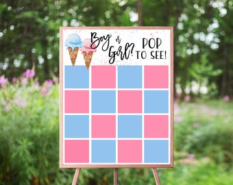 May include: A gender reveal sign on a wooden easel. The sign features two ice cream cones, one blue and one pink, with the text "Boy or Girl? Pop to See!" The sign has a grid of pink and blue squares.