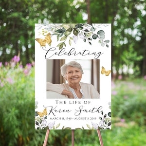 May include: A white memorial sign with a photo of a woman, green foliage, and gold butterflies. The text reads "Celebrating The Life of Karen Smith March 11, 1945 - August 5, 2019".
