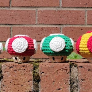 May include: Four colorful crochet mushroom figures in blue, red, green, and yellow with white accents. The mushrooms are arranged on a brick wall, showcasing their handmade details and vibrant colors. A whimsical, handcrafted item.