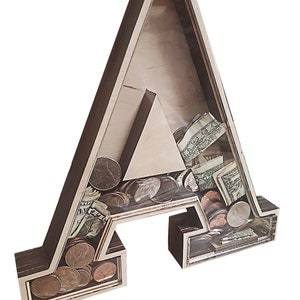 May include: A wooden letter A piggy bank with a clear acrylic front. The bank is filled with coins and dollar bills.