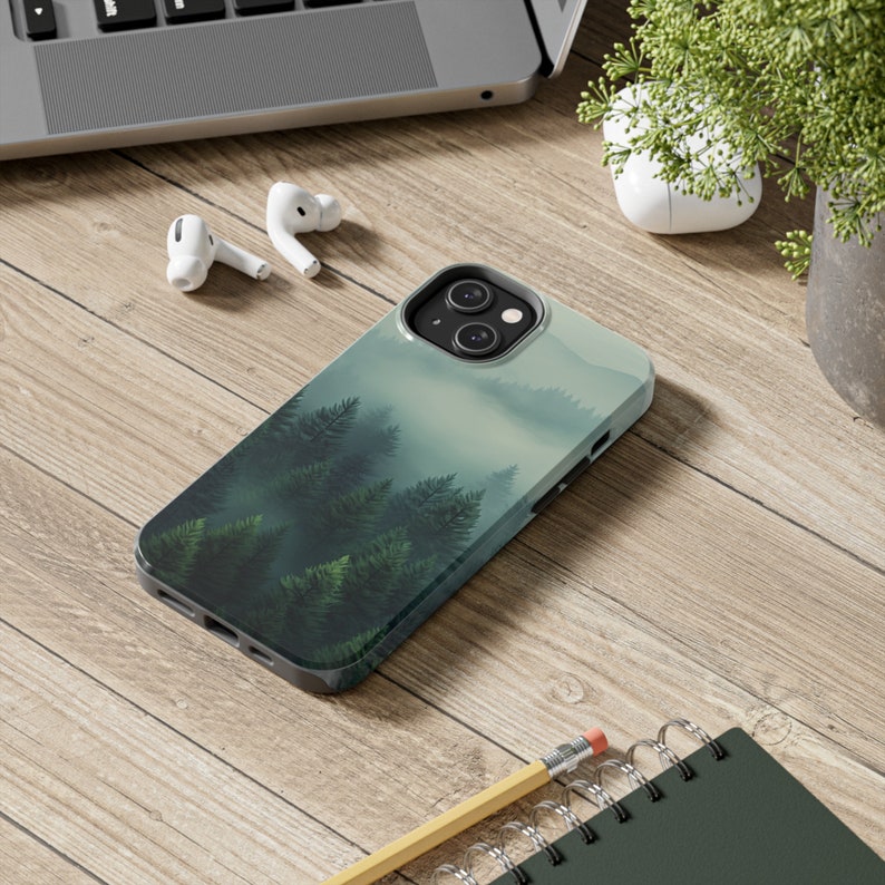 May include: A phone case with a misty forest scene. The case is black and has a green, blue, and white design. The phone case is on a wooden surface.