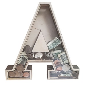 May include: Wooden letter A piggy bank with a clear plastic front. The bank is filled with coins and dollar bills.