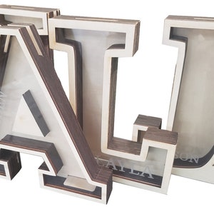 May include: Three wooden letter piggy banks, spelling out the name "ALI". Each letter is painted white with a brown wood grain finish. The letters are stacked on top of each other, with the "A" on the bottom, the "L" in the middle, and the "I" on top. The letters have a slot on the top for inserting coins. The letters are personalized with the names "ADENT", "KAYLA", and "SUN" on the front.