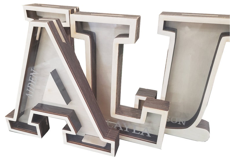 May include: Three wooden letter piggy banks, spelling out the name "ALI". Each letter is painted white with a brown wood grain finish. The letters are stacked on top of each other, with the "A" on the bottom, the "L" in the middle, and the "I" on top. The letters have a slot on the top for inserting coins. The letters are personalized with the names "ADENT", "KAYLA", and "SUN" on the front.