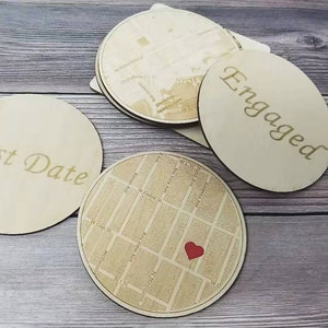 Può includere: Quattro sottobicchieri in legno con diversi design. Un sottobicchiere ha le parole "First Date" incise. Un altro sottobicchiere ha la parola "Engaged" incisa. Un terzo sottobicchiere ha una mappa di una città con un cuore rosso al centro. Il quarto sottobicchiere ha una mappa di una città.