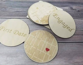May include: Four wooden coasters with different designs. One coaster has the words "First Date" engraved on it. Another coaster has the word "Engaged" engraved on it. A third coaster has a map of a city with a red heart in the centre. The fourth coaster has a map of a city.