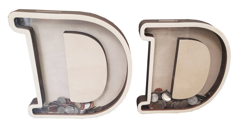 May include: Two wooden piggy banks shaped like the letter D. The piggy banks are made of light brown wood and have a clear plastic front. The piggy bank on the left is empty, while the piggy bank on the right is filled with coins.