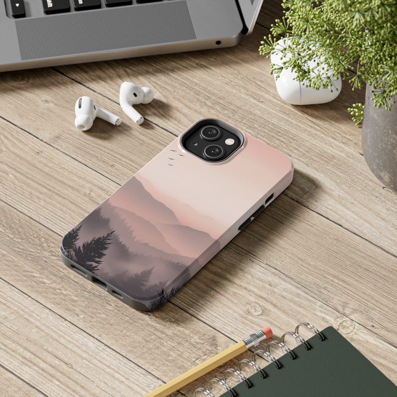 May include: A pink and gray phone case with a mountain range and a sunset in the background. The case has a raised camera bump and a black trim.