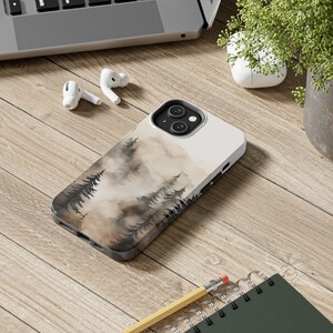 May include: A white phone case with a watercolor design of a foggy forest with brown and black trees. The phone case is on a wooden surface.