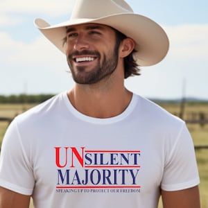 Könnte beinhalten: Ein weißes T-Shirt mit dem Text "UNSILENT MAJORITY" in Rot und Blau. Das T-Shirt wird von einer Person getragen, die einen Cowboyhut trägt und vor einem verschwommenen Hintergrund im Freien lächelt.
