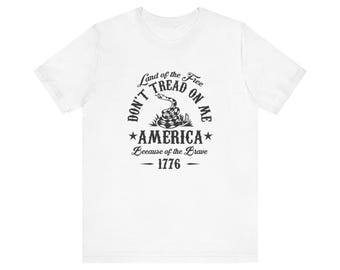 May include: White t-shirt with a black graphic. The design includes a coiled snake and the text "Don't Tread on Me" and "America Because of the Brave 1776".