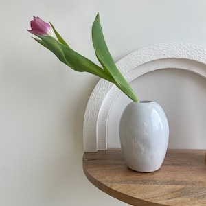 May include: A white ceramic vase holds a single pink and purple tulip with green leaves. The vase sits on a wooden shelf with a white, textured, arched wall decoration in the background. The overall aesthetic is minimalist and modern.