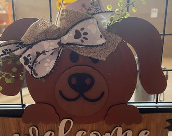 May include: A brown wooden dog-shaped welcome sign with a burlap bow and a white and brown paw print ribbon. The sign is hanging on a wire fence.
