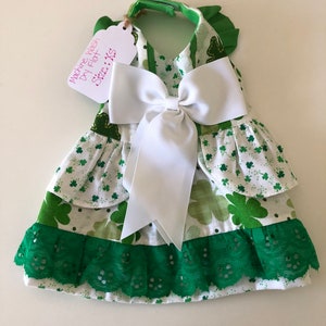 May include: A green and white dog dress with a white bow and green lace trim. The dress has a shamrock pattern and says "Machine Wash Dry Flat Size: XS".
