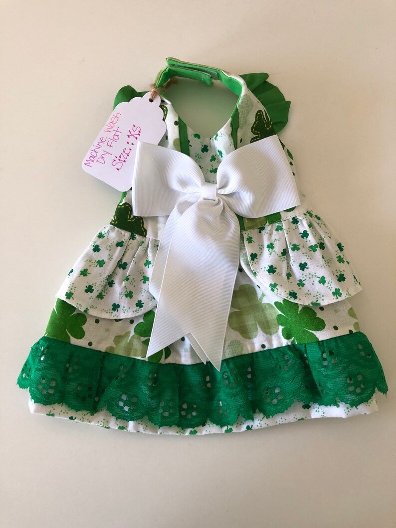 May include: A green and white dog dress with a white bow and green lace trim. The dress has a shamrock pattern and says "Machine Wash Dry Flat Size: XS".