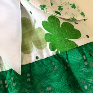 May include: A white fabric with green shamrock print and green lace trim. The fabric has a white ribbon attached to it.