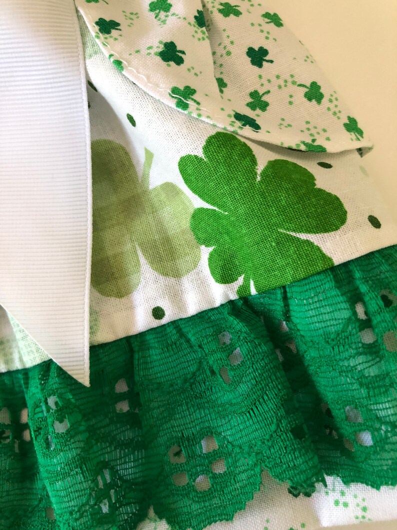 May include: A white fabric with green shamrock print and green lace trim. The fabric has a white ribbon attached to it.