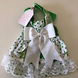 May include: Green and white dog dress with a white bow and lace trim. The dress has a shamrock pattern and says "Martine Roos Dog Frock Size: XS".