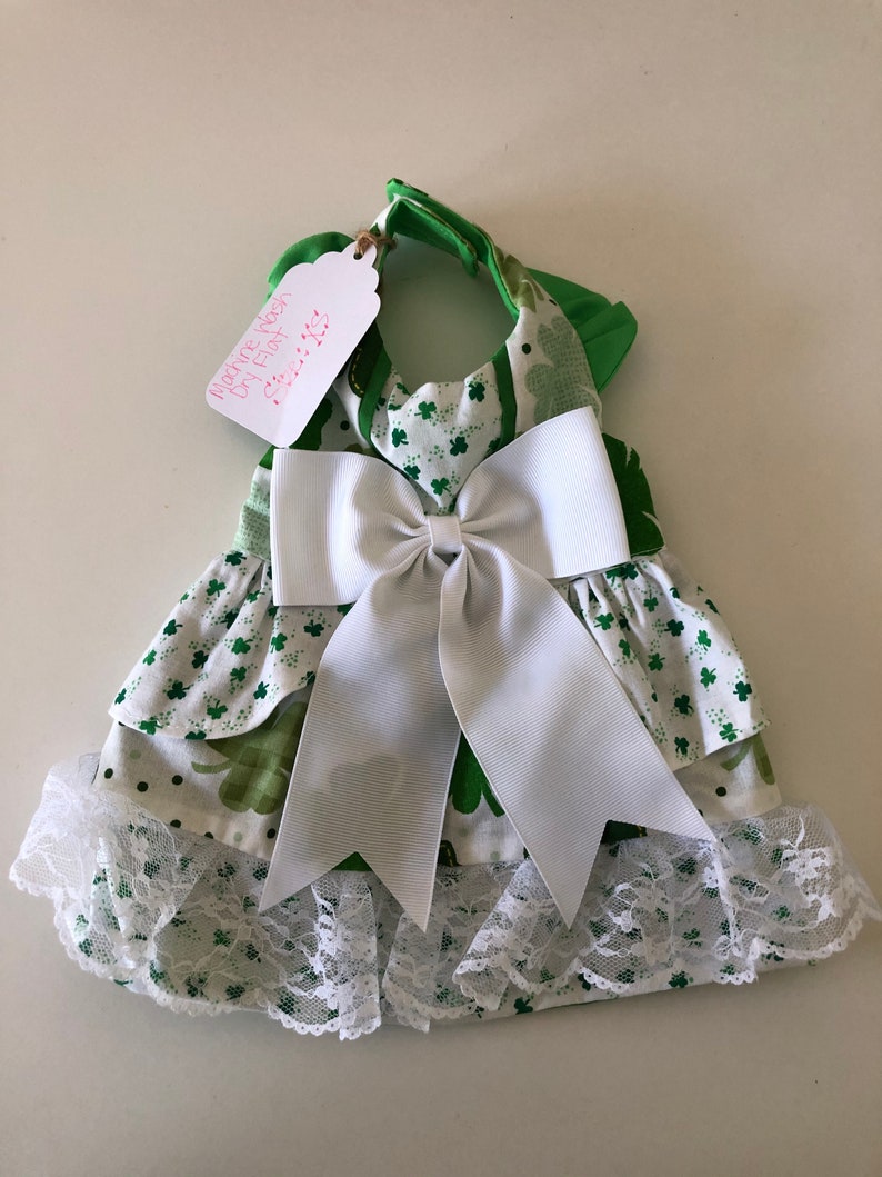 May include: Green and white dog dress with a white bow and lace trim. The dress has a shamrock pattern and says "Martine Roos Dog Frock Size: XS".