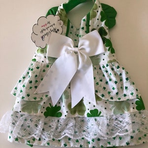 May include: A green and white dog dress with a shamrock print and a white bow. The dress has a ruffled skirt and a lace trim. The tag says "Happy St. Patrick's Day! You are my lucky charm!"