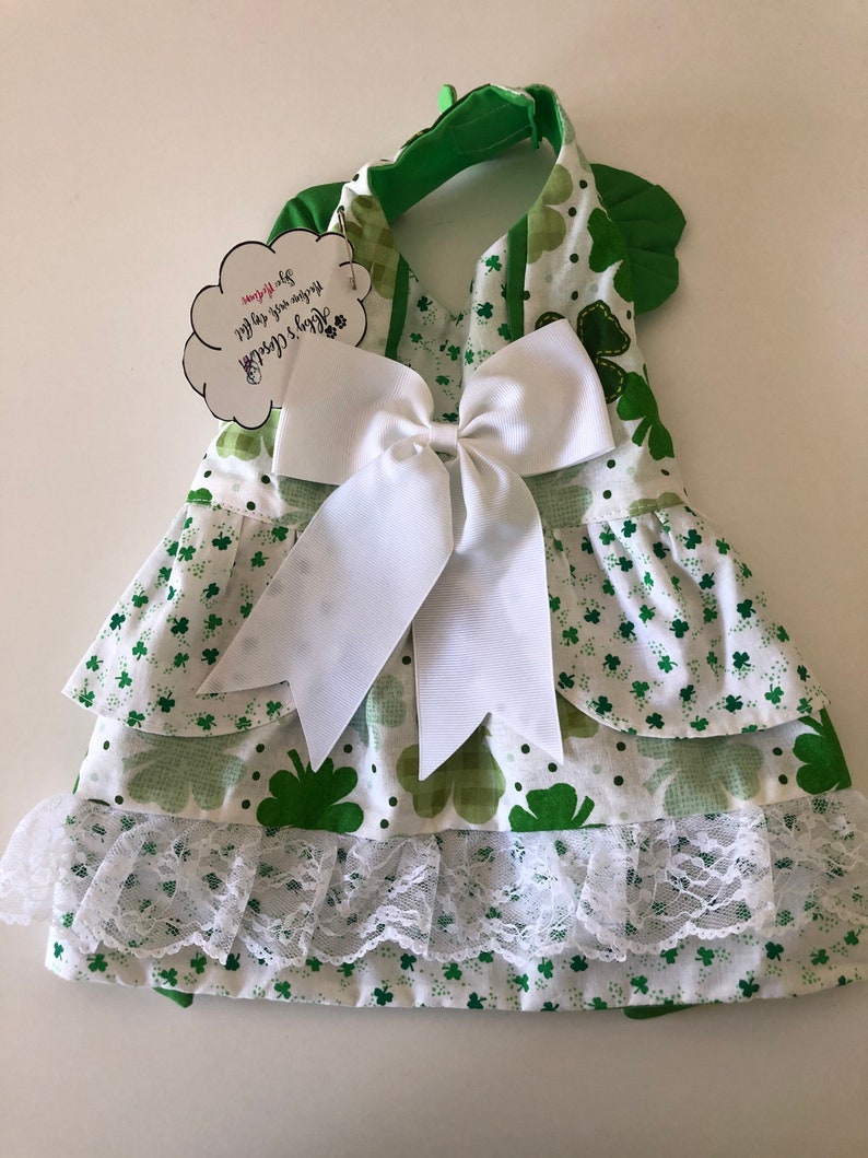 May include: A green and white dog dress with a shamrock print and a white bow. The dress has a ruffled skirt and a lace trim. The tag says "Happy St. Patrick's Day! You are my lucky charm!"