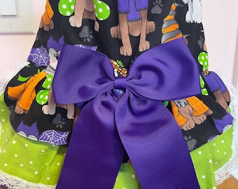 May include: A Halloween-themed dog dress with a black background featuring dog illustrations, paw prints, and spiderwebs. The dress has a large purple bow, green polka dot trim, and white lace edging. The dress has the word "SUPER" on it.