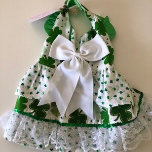 May include: A green and white dress with a shamrock print and a white bow. The dress has a lace trim and a green sequin trim.