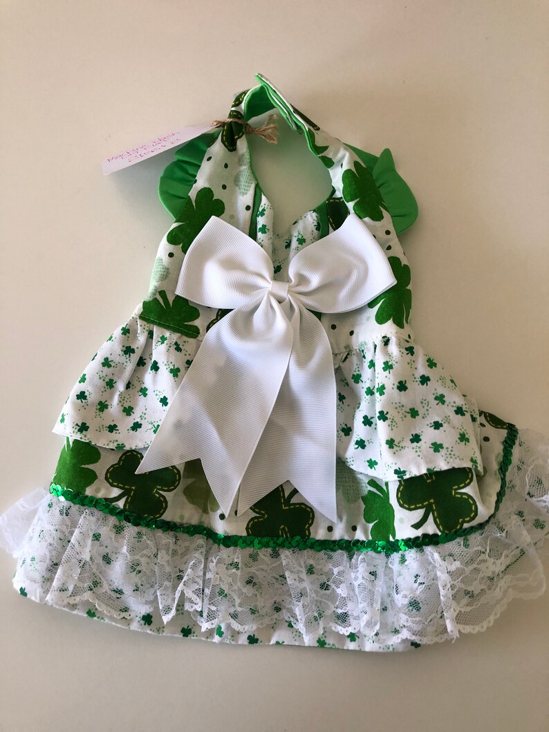 May include: A green and white dress with a shamrock print and a white bow. The dress has a lace trim and a green sequin trim.
