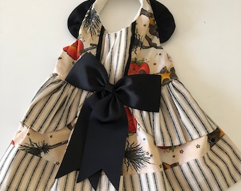 May include: A black and white striped dog dress with a black bow and a Halloween-themed print featuring witches, brooms, and stars. The dress has a halter neckline and a ruffled skirt.