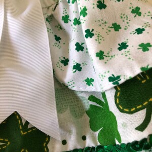 May include: A white dress with a green shamrock print and a white bow. The dress has a green sequin trim and a white lace ruffle.