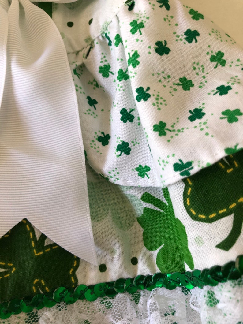 May include: A white dress with a green shamrock print and a white bow. The dress has a green sequin trim and a white lace ruffle.