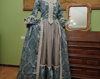 May include: A teal and gray brocade dress with a lace trim and a fitted bodice. The dress is on a mannequin and has a long skirt.