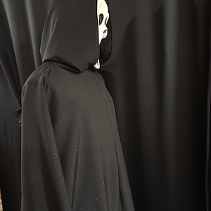 Könnte beinhalten: Ein schwarzer Kapuzenmantel mit einer weißen Maske. Die Maske hat ein bleiches, schädelähnliches Aussehen mit dunklen Augenhöhlen und einem klaffenden Mund. Der Umhang besteht aus einem dunklen, fließenden Stoff.