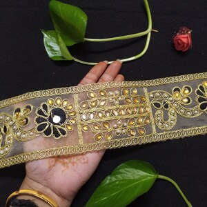 May include: Gold and black embroidered trim with mirror embellishments and pearl beads. The trim is on a sheer fabric.