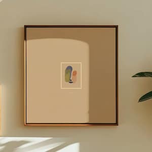 May include: A square framed wall art piece featuring a small abstract face illustration on a tan background, displayed alongside a smaller framed landscape print, a white textured armchair with a red pillow, and a round red floor cushion.