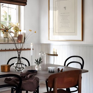 May include: A dining room with a white wainscot wall, a framed print with the text "The sampler of love", a wooden table with black chairs, a glass vase with dried flowers, and a candle on the table.