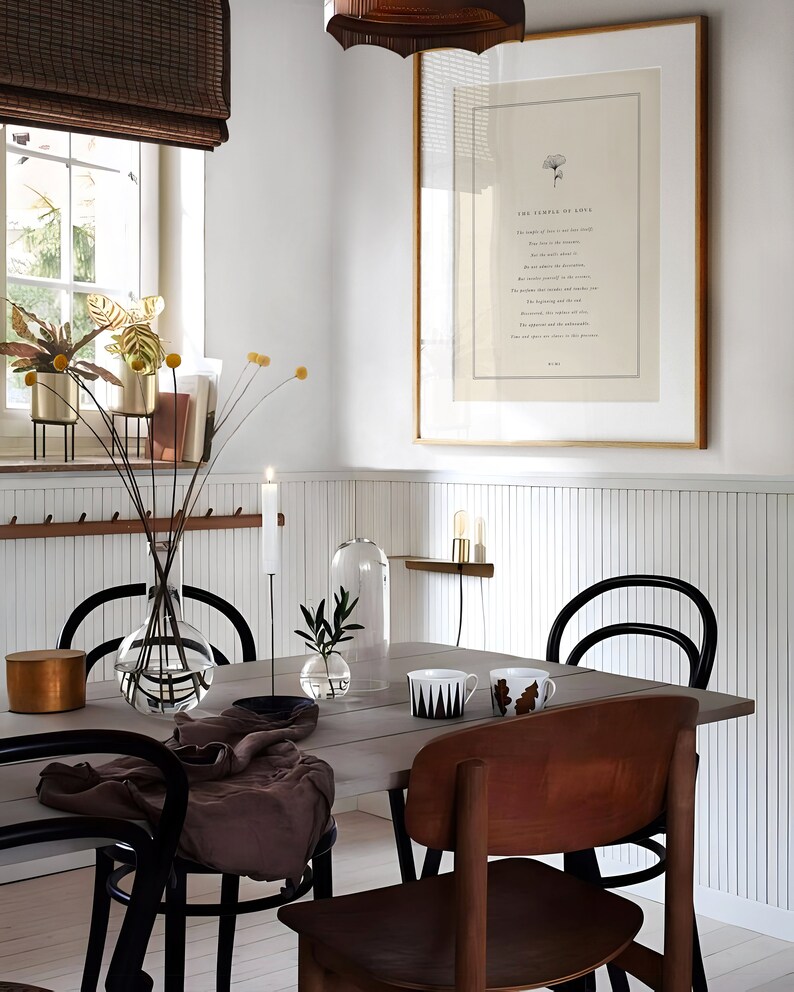 May include: A dining room with a white wainscot wall, a framed print with the text "The sampler of love", a wooden table with black chairs, a glass vase with dried flowers, and a candle on the table.