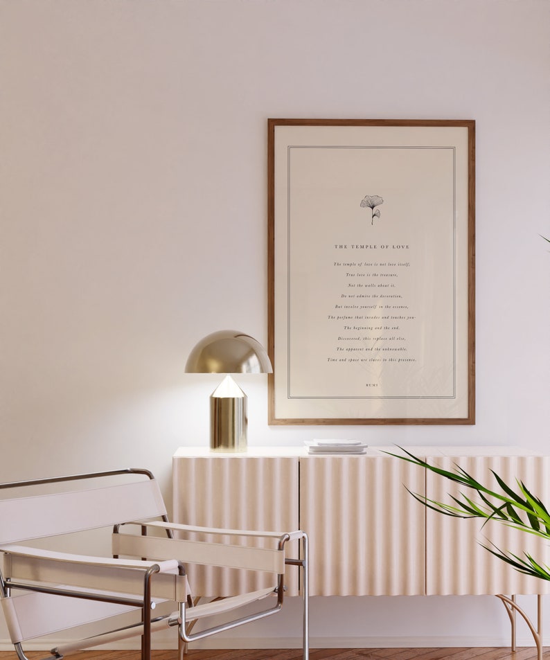 May include: A framed print with a white background and black text. The text reads "The Temple of Love" and includes a poem about love. The print is hanging on a white wall above a light brown cabinet with a gold lamp and a white chair.