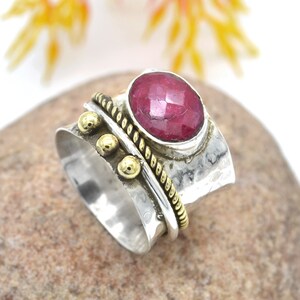 May include: A silver and gold ring with a red ruby gemstone. The ring has a hammered texture and a band with a rope design and gold beads.