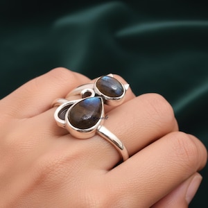May include: Two sterling silver rings with teardrop-shaped labradorite stones. The stones display a mix of blue, brown, and gray hues. The rings are on a hand, showcasing the jewelry's design and color.