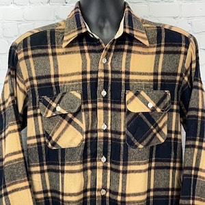 May include: A button-down flannel shirt in a plaid pattern of tan, navy blue, and grey. The shirt has two chest pockets with button closures and a white collar. The long-sleeved shirt is displayed on a black mannequin.
