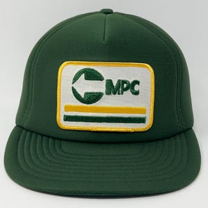 Puede incluir: Una gorra de béisbol de color verde oscuro con un parche rectangular en la parte delantera. El parche tiene un fondo blanco con borde amarillo, mostrando un logotipo circular verde, el texto MPC y dos franjas horizontales en amarillo y verde.