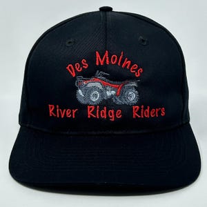 Könnte beinhalten: Schwarze Baseballkappe mit roter Stickerei "Des Moines River Ridge Riders". Das Design beinhaltet eine rote und graue ATV-Grafik. Die Kappe hat einen gebogenen Schirm und einen Knopf oben.