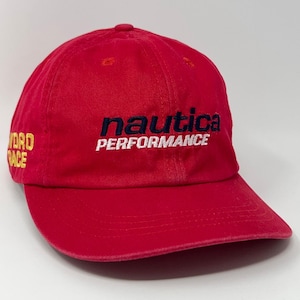 Puede incluir: Gorra de béisbol roja Nautica con la palabra "nautica" en azul marino y "PERFORMANCE" en blanco bordado en la parte delantera. El lateral de la gorra muestra el texto "WORD RACE". El sombrero está hecho de una tela suave.