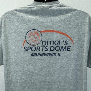Pode incluir: Camiseta cinza mesclada com o logotipo Ditka's Sports Dome. O logotipo inclui um rosto de desenho animado dentro de um círculo, com as palavras "DITKA'S SPORTS DOME" e "BOLINGBROOK, IL" abaixo.